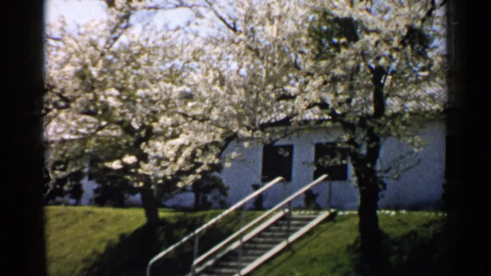 A 1952 Japan 8mm frame of a white building seen through trees, with exterior railings, stairs, and a sloped green lawn.