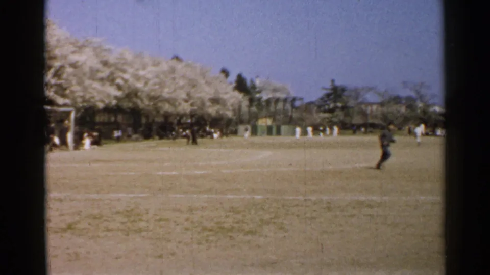 Wide 1952 Japan park view with an open playing field, distant people, a goal frame, and blossom trees along the background.