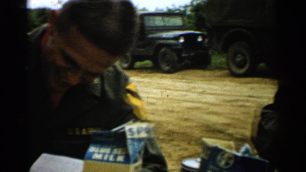A uniformed man writes or checks notes outdoors, with dark military vehicles behind him in Japan 1952 footage.