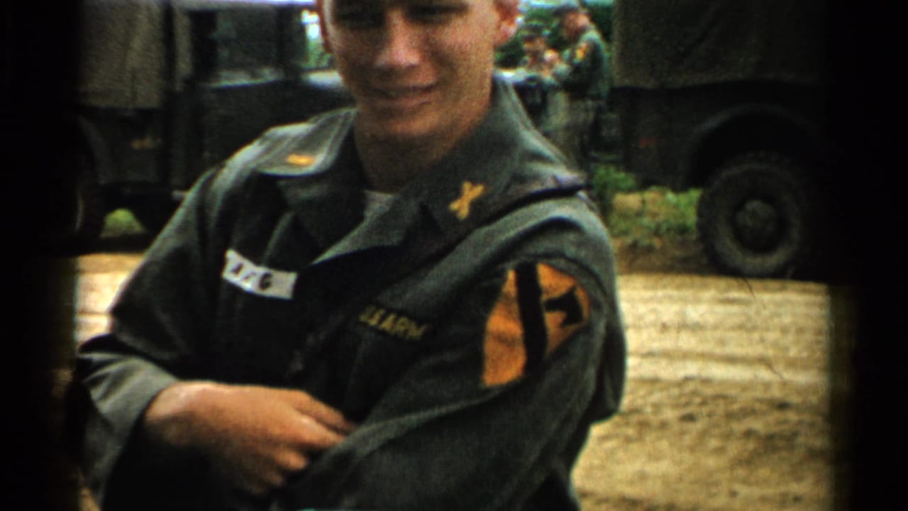 Young soldier in green uniform smiles near military trucks and other soldiers.