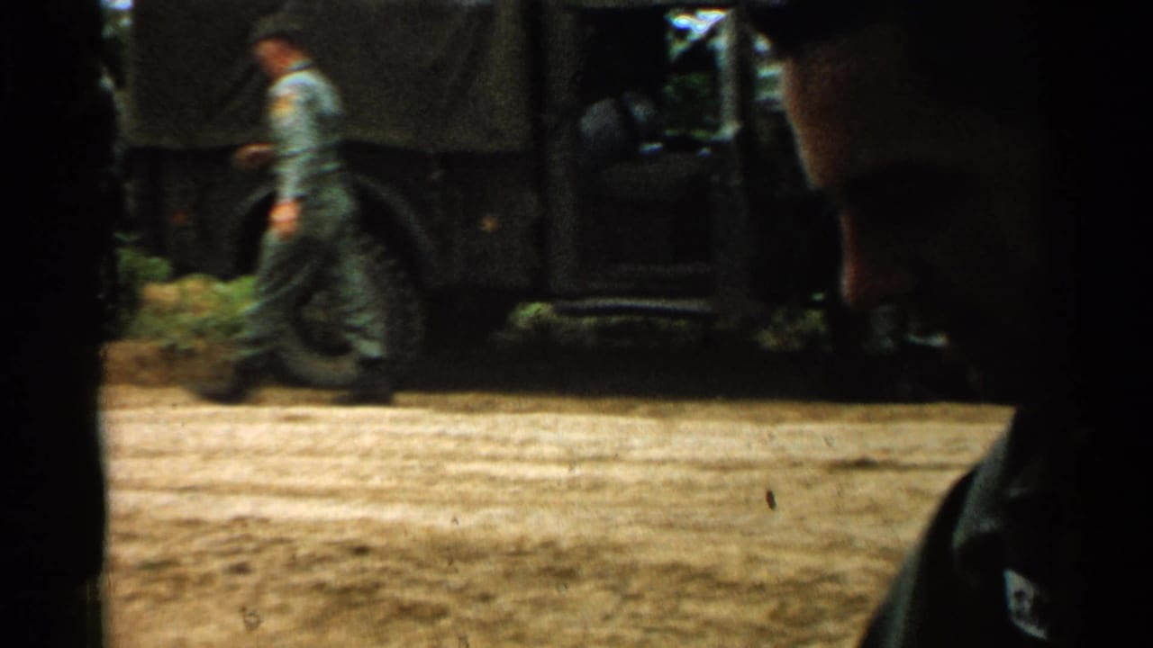 In the 1952 Japan military scene, a uniformed soldier walks near a dark truck while the camera catches a partial foreground figure.