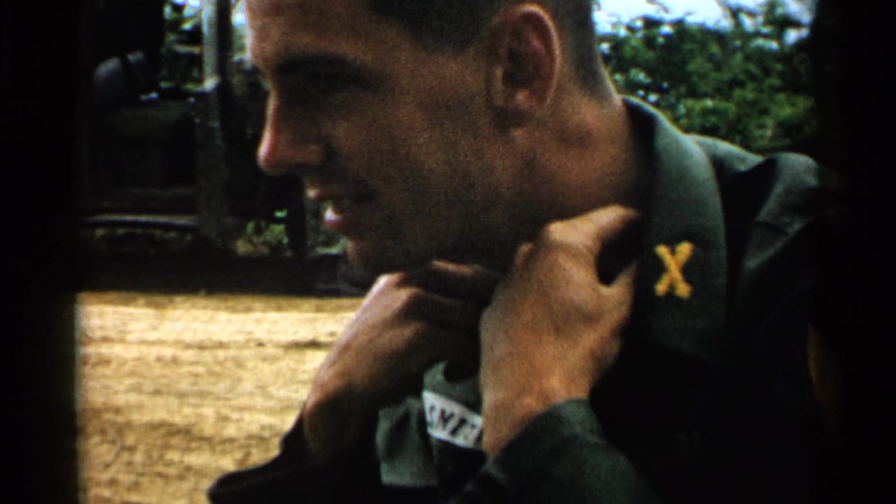 A young American soldier in 1952 Japan adjusts his collar in profile, with a truck and outdoor staging area behind him.