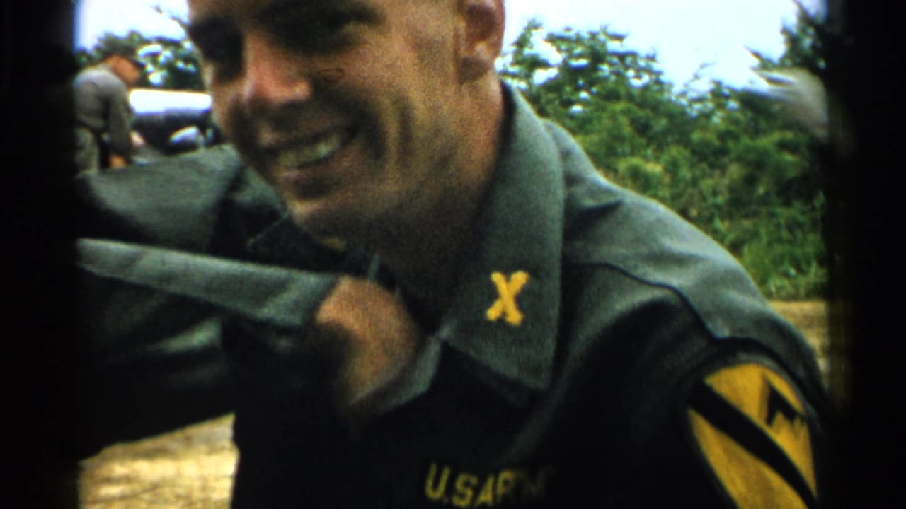 In a 1952 Japan home-movie frame, the soldier smiles while pulling at his uniform collar outdoors.