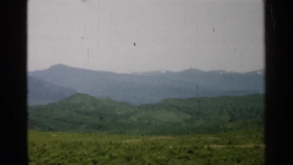 1952 Japan 8mm view of rolling green hills before a hazy mountain range with small snow patches, framed by dark window-like edges.