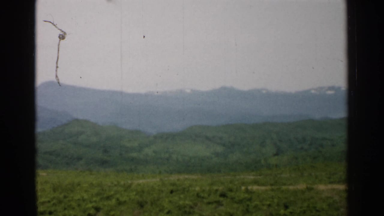 1952 Japan landscape frame with green foothills, a blue-gray mountain ridge, faint snow, and visible 8mm dust and scratches.