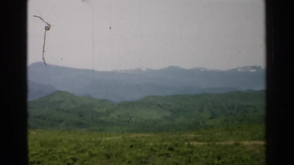 1952 Japan landscape frame with green foothills, a blue-gray mountain ridge, faint snow, and visible 8mm dust and scratches.