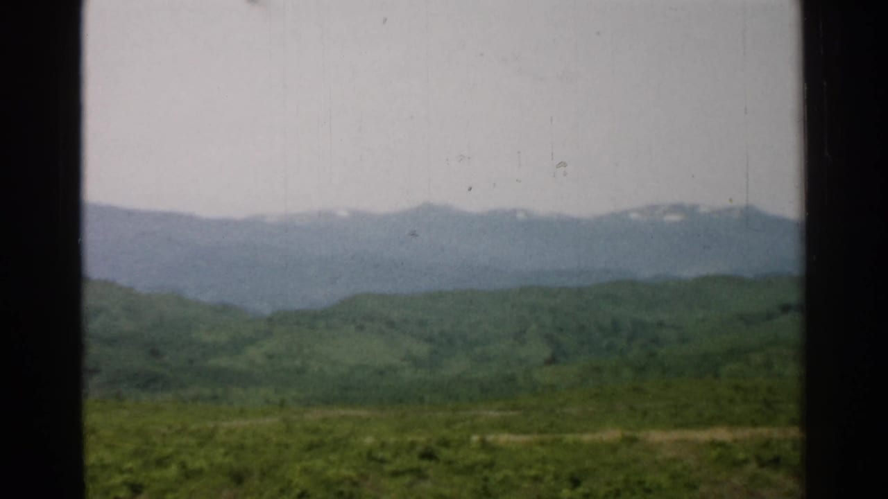 Fixed 8mm home-movie view in Japan, showing a green valley foreground and distant snow-patched mountains beneath an overcast sky.