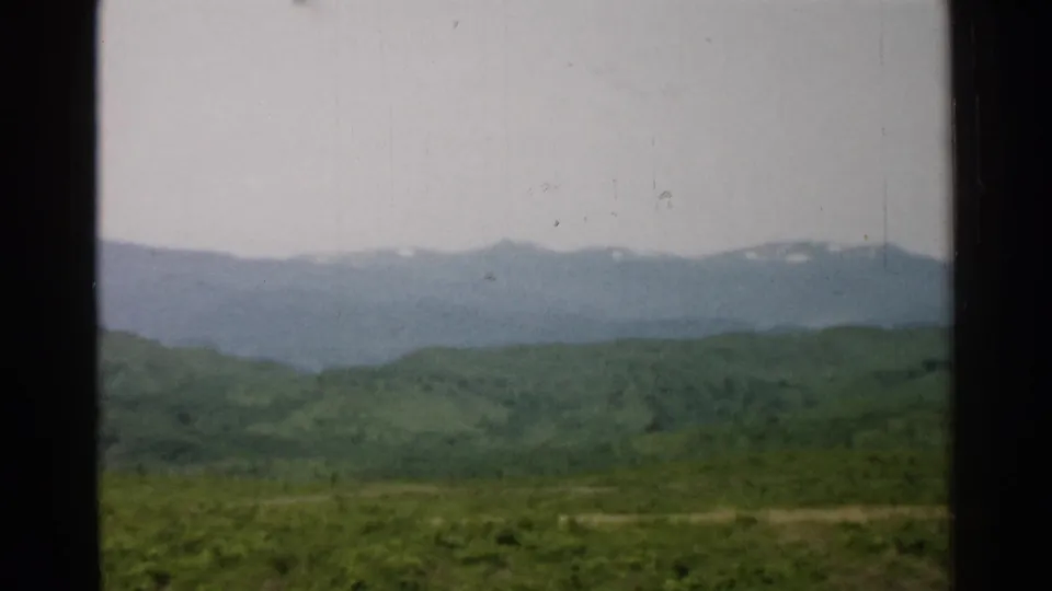Fixed 8mm home-movie view in Japan, showing a green valley foreground and distant snow-patched mountains beneath an overcast sky.
