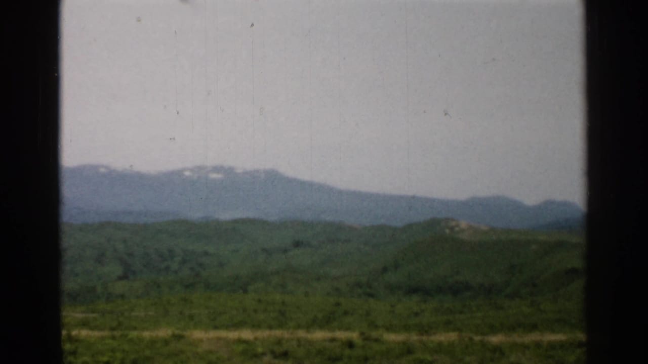1952 Japan travel footage frame with layered green hills, a distant hazy ridge, small snow remnants, and dark film or window borders.