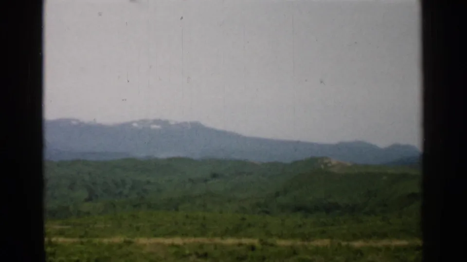 1952 Japan travel footage frame with layered green hills, a distant hazy ridge, small snow remnants, and dark film or window borders.