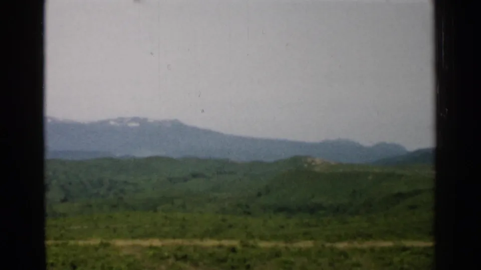 1952 Japan home-movie frame showing green hills, distant mountains with small snow patches, and a bright pale sky.