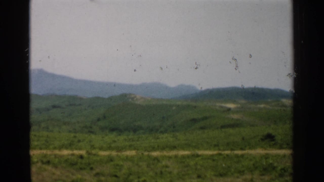 1952 Japan countryside with layered green hills, a light dirt road, distant mountains, and dark film or window borders.