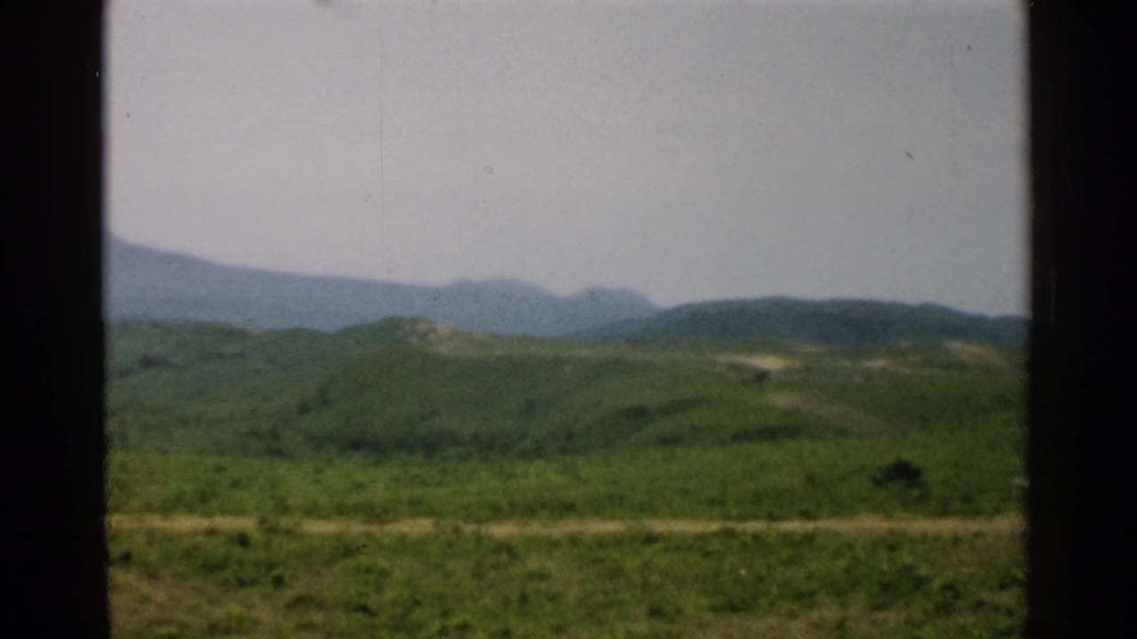 Soft 8mm frame of 1952 Japan countryside, showing green hills, a pale road, and hazy mountains through a window-like foreground.
