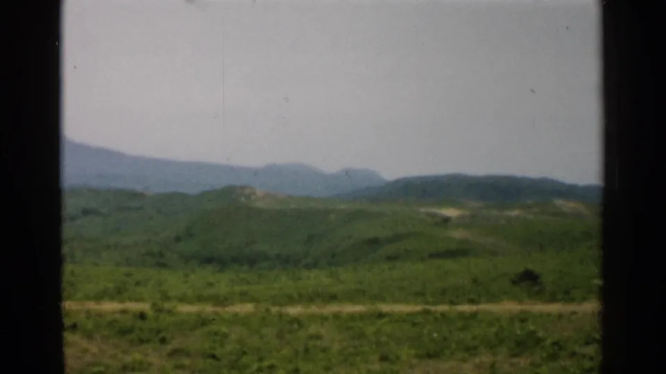 Soft 8mm frame of 1952 Japan countryside, showing green hills, a pale road, and hazy mountains through a window-like foreground.