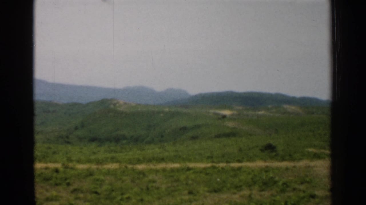 1952 Japan rural landscape with rolling green terrain, a light dirt road, distant mountains, and visible 8mm film texture.