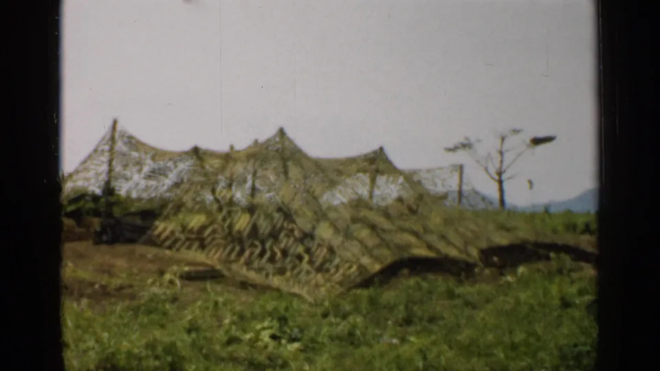 1952 Japan 8mm frame showing a low camouflage net tent in grass, with a bare tree, poles, pale sky, and distant mountains.