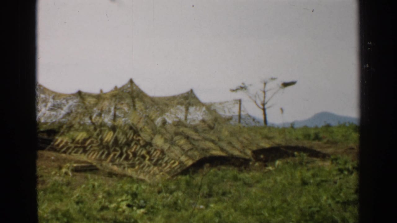 1952 Japan 8mm frame of a camouflaged low tent or shelter on a grassy slope, with sparse trees, poles, and mountains beyond.