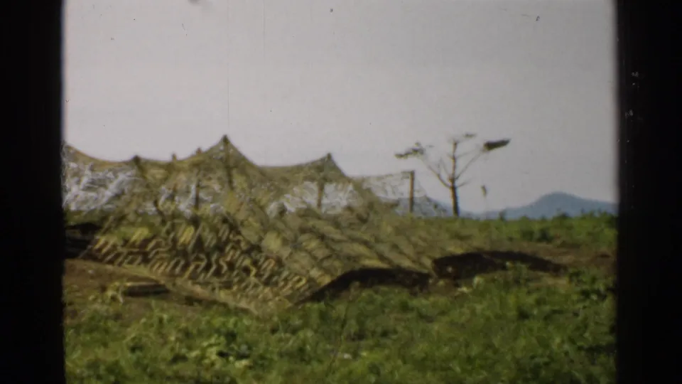 1952 Japan 8mm frame of a camouflaged low tent or shelter on a grassy slope, with sparse trees, poles, and mountains beyond.
