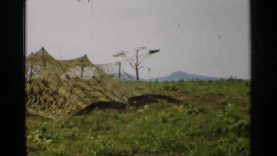 1952 Japan 8mm frame showing camouflage netting near trench-like earthworks in grass, with a lone bare tree and low mountains beyond.