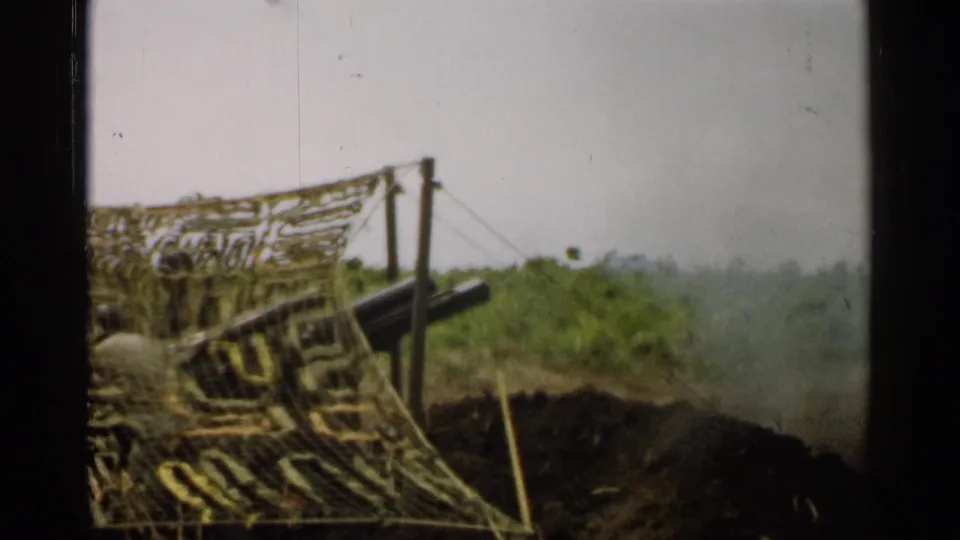1952 Japan 8mm frame of artillery behind camouflage netting, with foliage and a faint smoke plume near the field position.