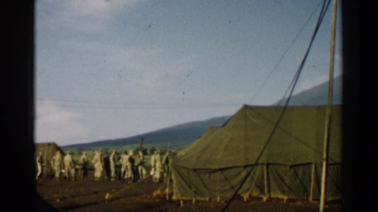Canvas military tents with soldiers standing nearby on dirt ground below distant mountains.