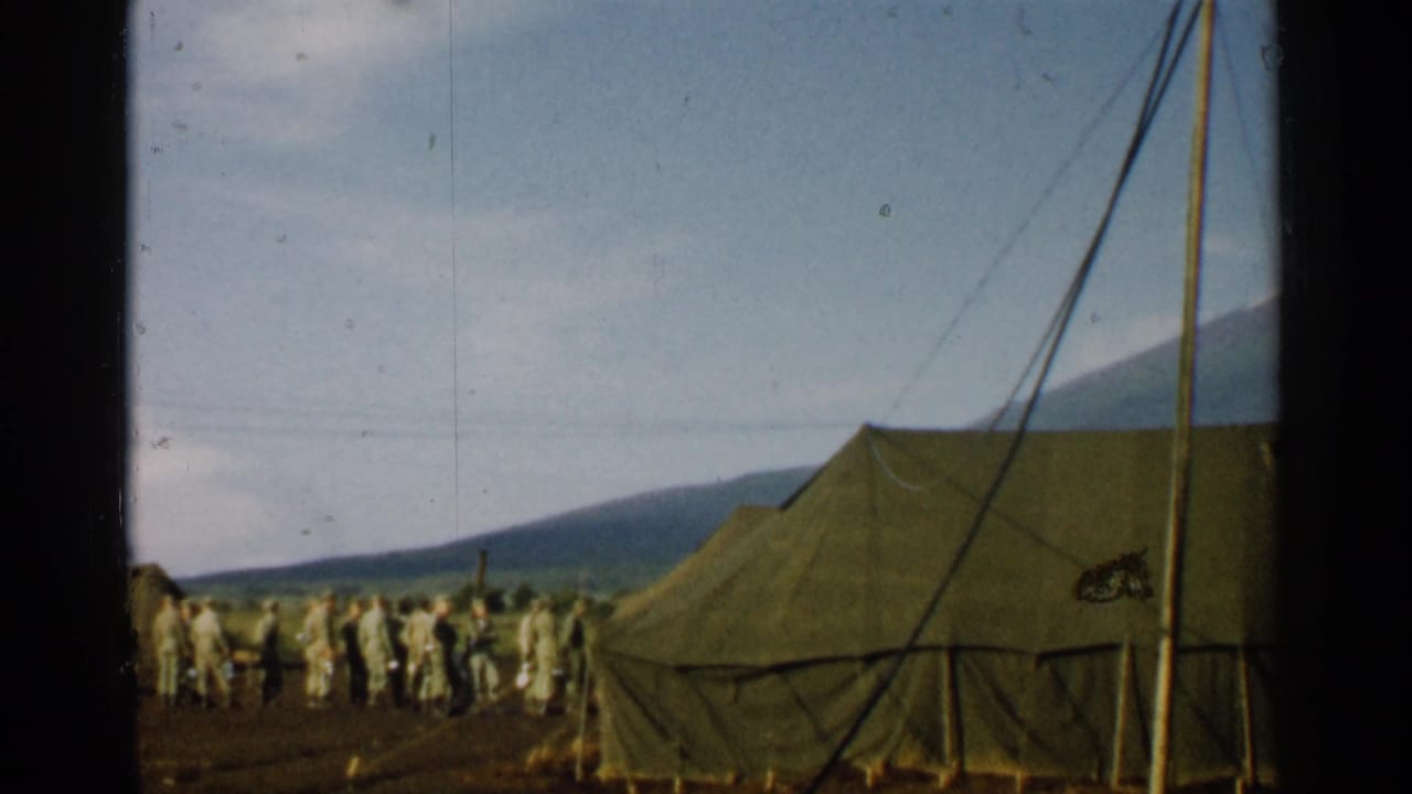 1952 Japan 8mm frame with soldiers near olive drab tents, mountain backdrop, tent rigging, and a partly obscured camouflaged object.