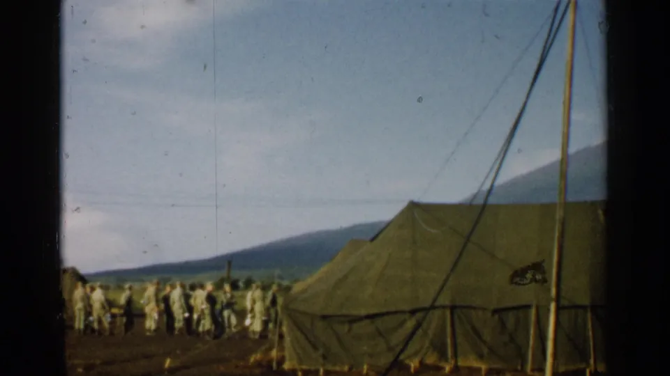 1952 Japan 8mm frame with soldiers near olive drab tents, mountain backdrop, tent rigging, and a partly obscured camouflaged object.