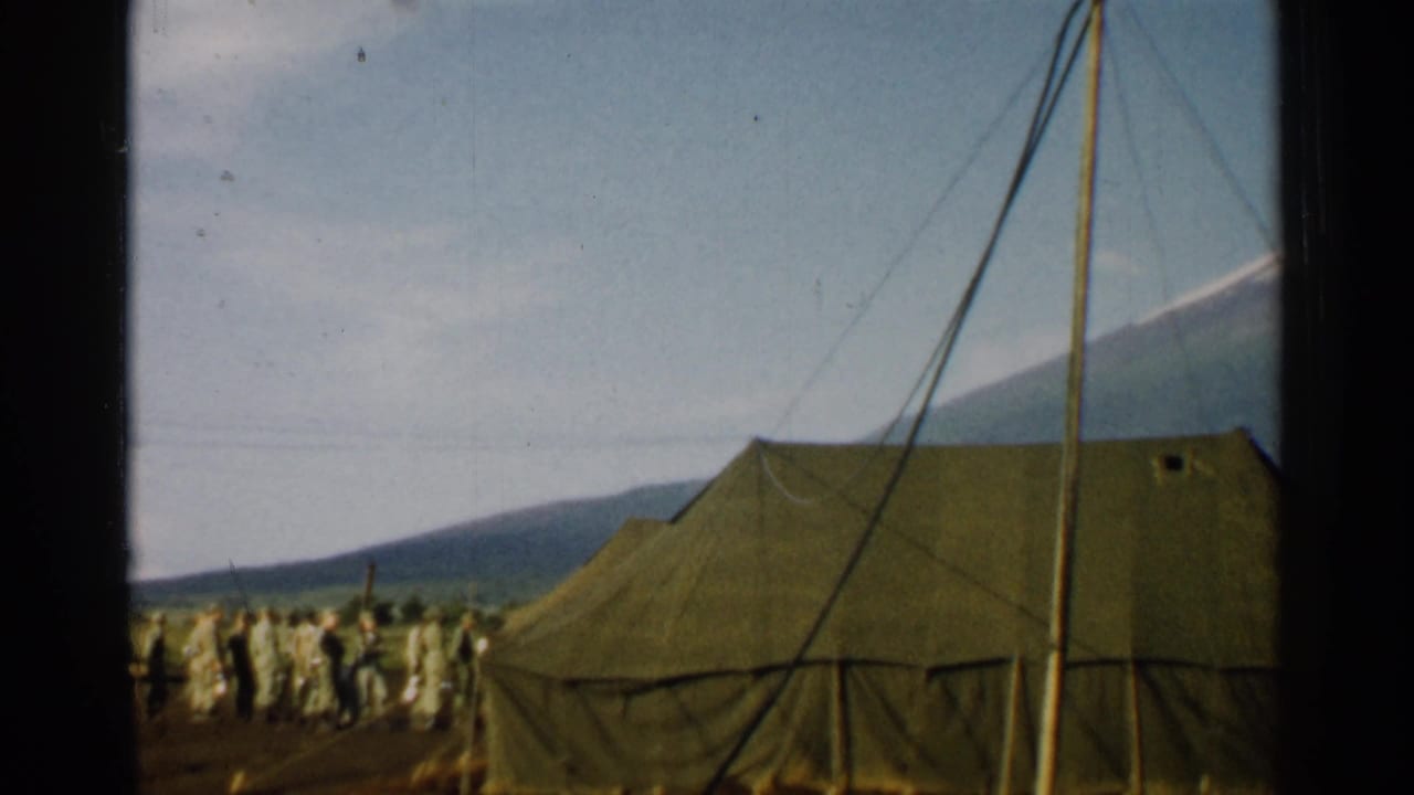 1952 Japan 8mm frame showing a close view of a canvas army tent, soldiers clustered at left, guy lines, and a mountain rising behind camp.
