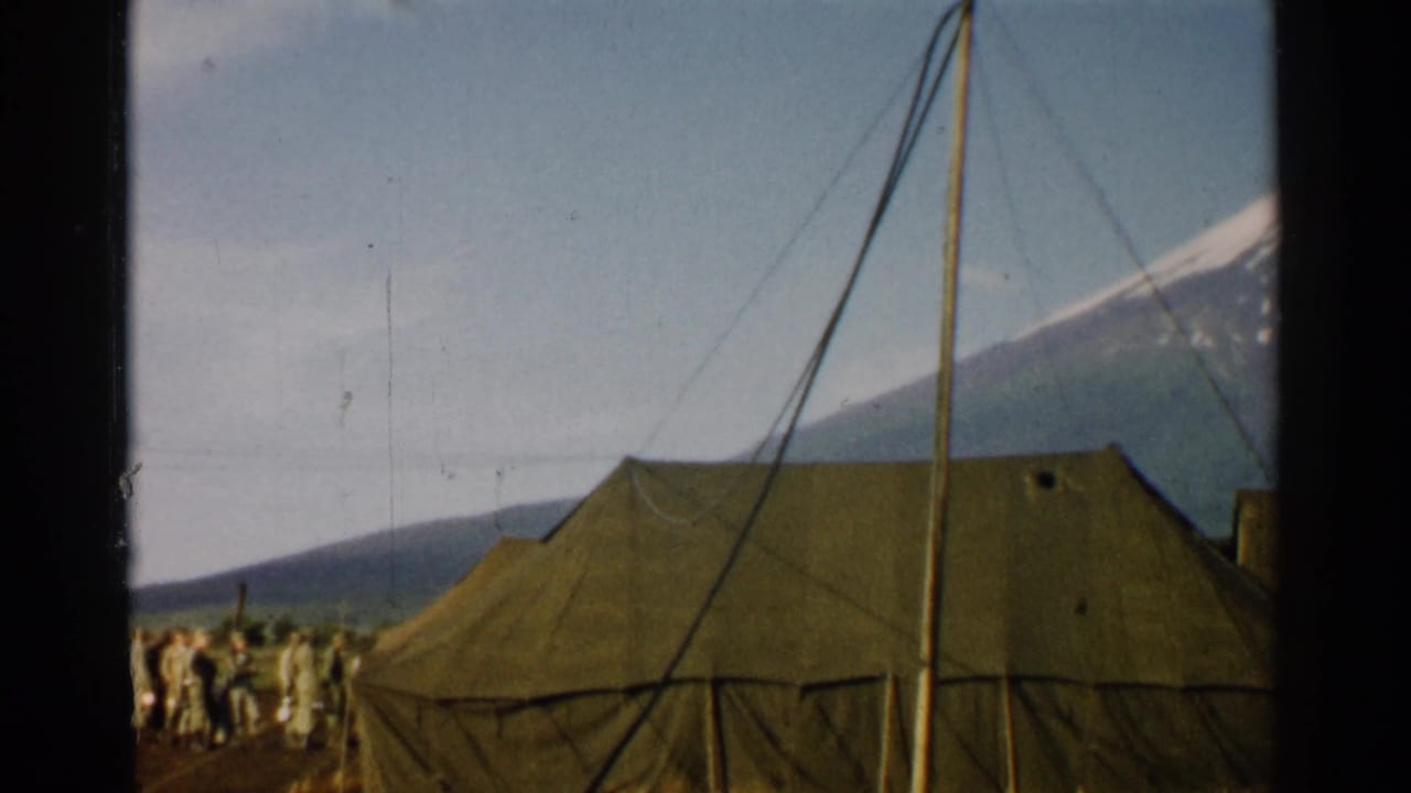 1952 Japan 8mm frame centered on an olive drab tent, with soldiers partly visible at left, rigging lines, dirt ground, and mountains beyond.