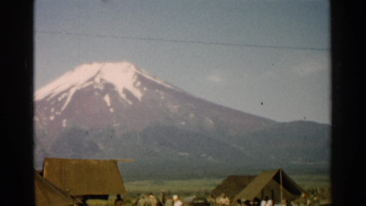 1952 Japan home-movie frame of Mount Fuji over a rural settlement, with thatched structures and small figures near the foreground.