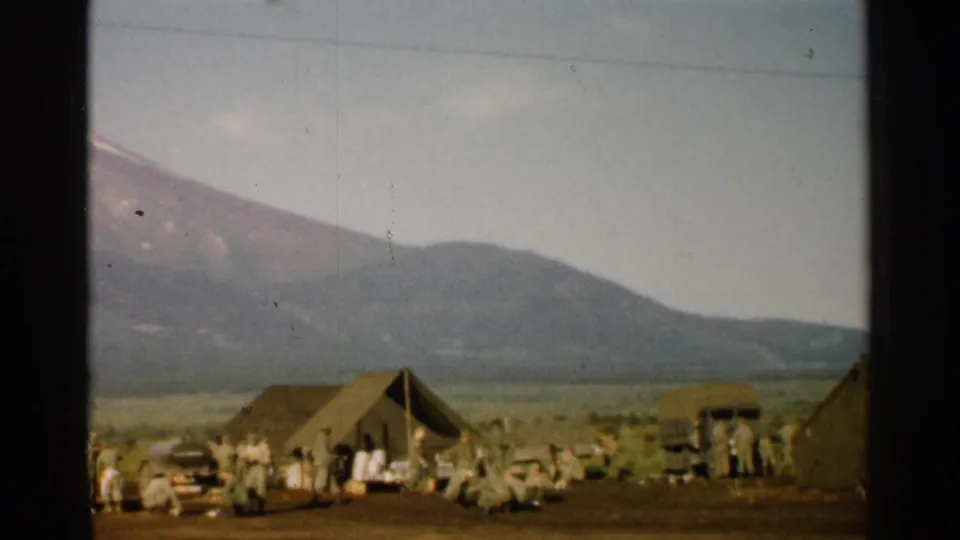 1952 Japan frame showing a military-style camp with tents, gathered personnel, and a covered vehicle under Mount Fuji's foothills.