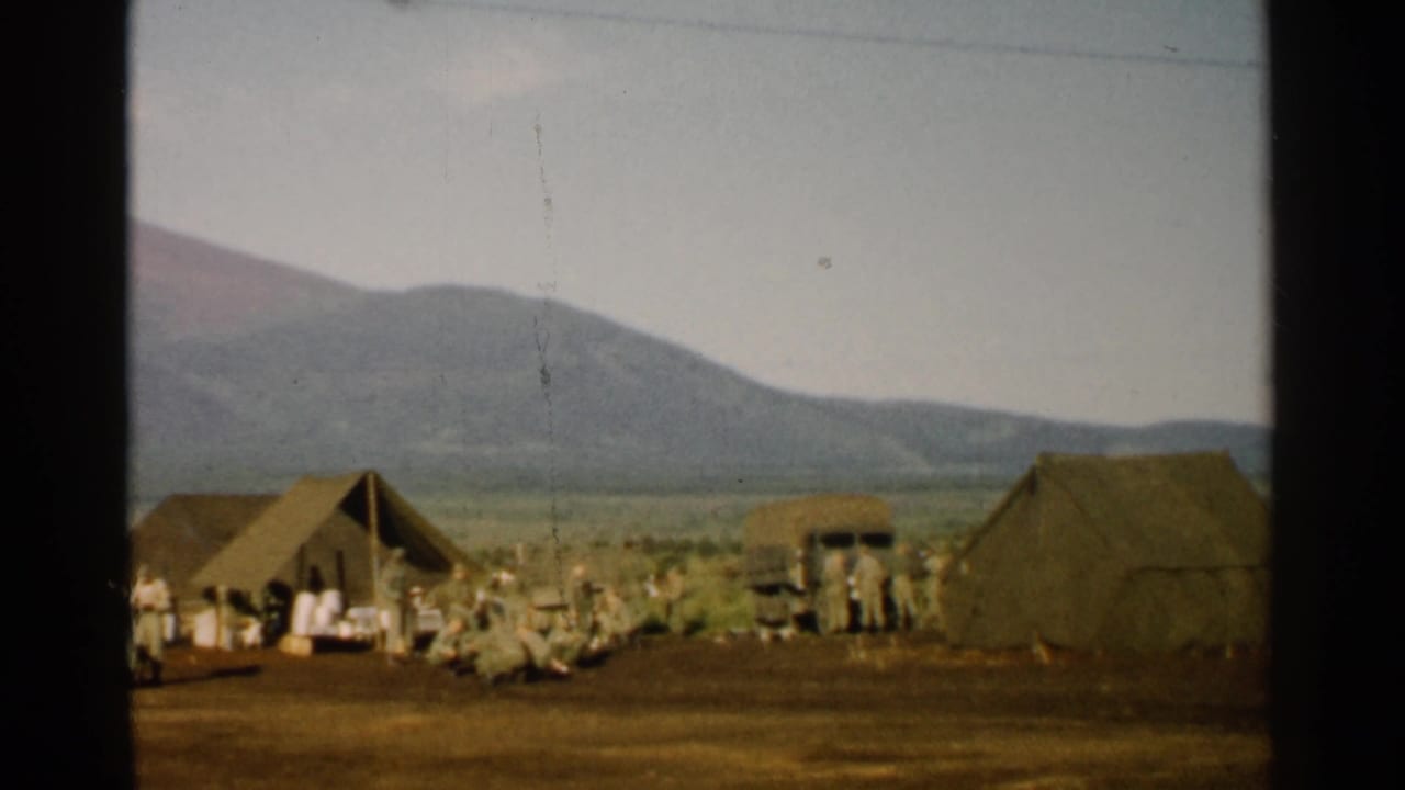 Soldiers stand near tents and a truck in a field below mountains.