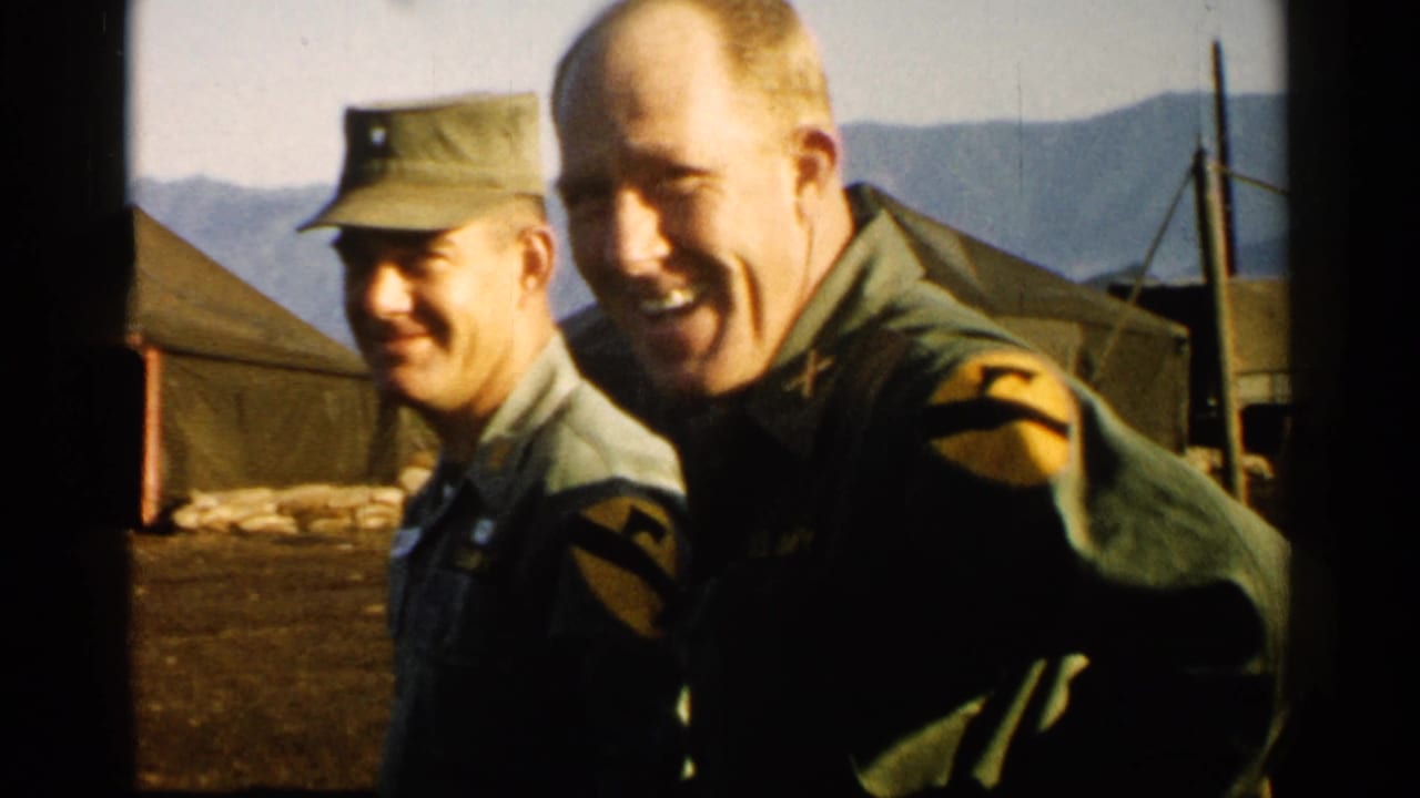 Two uniformed soldiers smile near tents with mountains behind them.