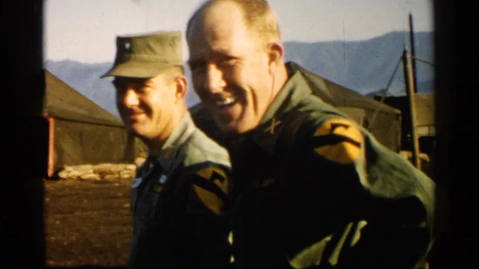 1952 Japan home-movie frame of two U.S. Army-uniformed men smiling at the camera beside canvas tents.