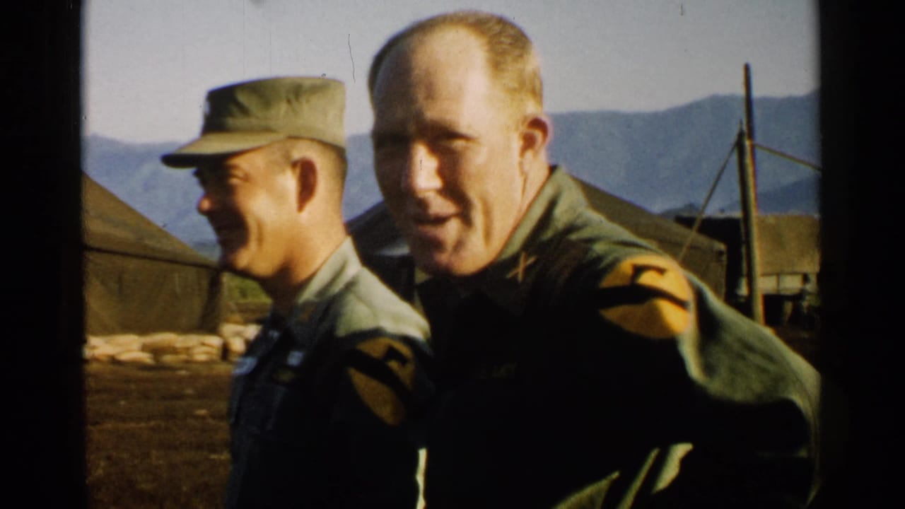 Two U.S. Army officers stand outdoors near tents and mountains.
