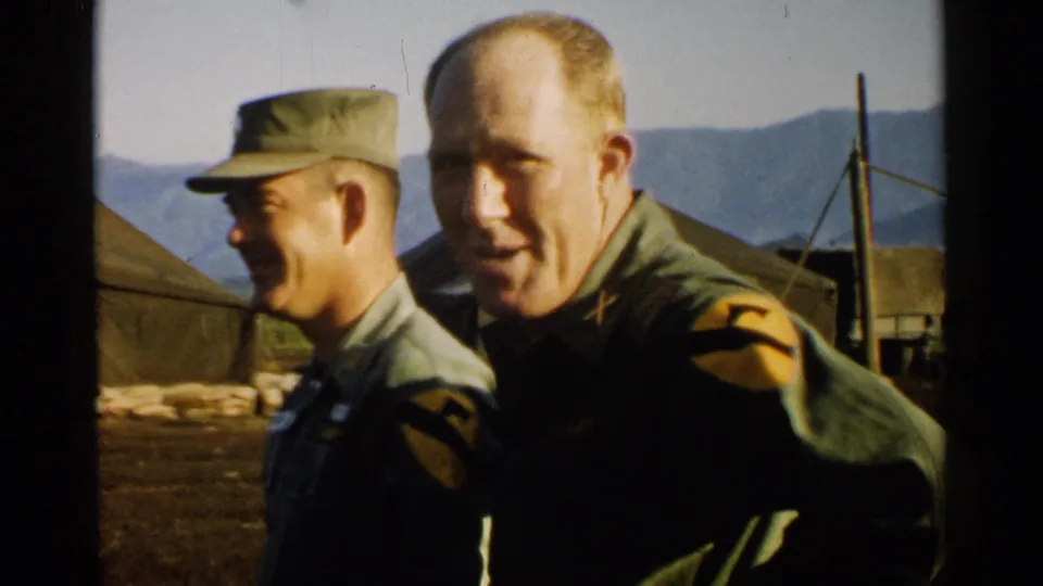 Two U.S. Army officers in 1952 Japan stand close to the camera at an outdoor camp with tents and mountains behind them.