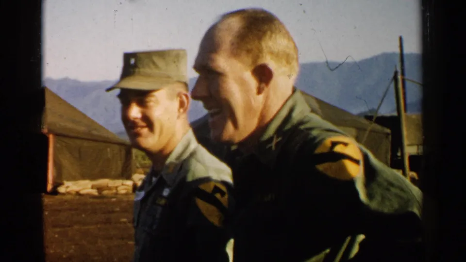 Two U.S. Army officers in Japan appear in profile at a camp, with canvas tents, sandbags, poles, and mountains in the background.