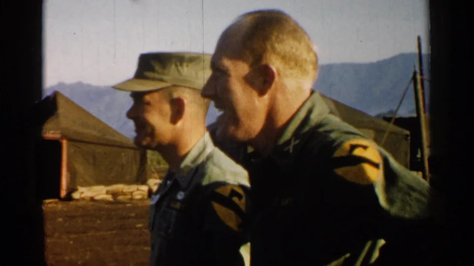 Two U.S. Army officers at the Japan camp turn left as the handheld 8mm camera frames tents, sandbags, and mountain scenery.