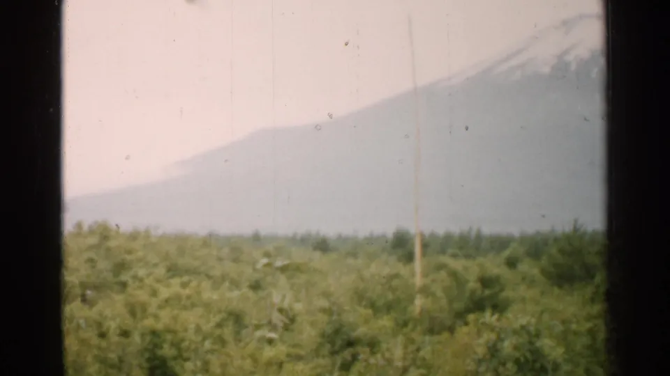 1952 Japan 8mm frame showing a snow-capped mountain partly obscured by haze above dense green forest.