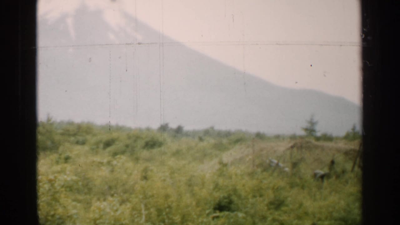 1952 Japan 8mm frame showing a hazy mountain beyond green grassland, seen through a dark window frame.