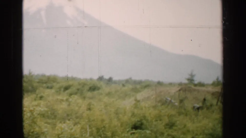 1952 Japan 8mm frame showing a hazy mountain beyond green grassland, seen through a dark window frame.