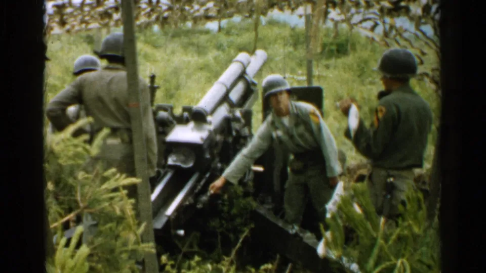 1952 Japan archival frame with soldiers in helmets around a field cannon, framed by foliage and dark edges from the 8mm scan.