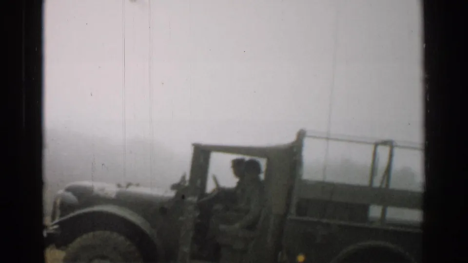 A soft 1952 Japan 8mm frame shows a canvas-top military vehicle near the camera, with roadside structures faintly visible behind it.