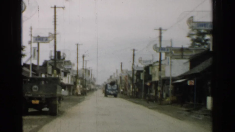 Forward-looking 1952 Japan street view with a military truck ahead, low buildings, utility poles, signage, and another vehicle in the distance.