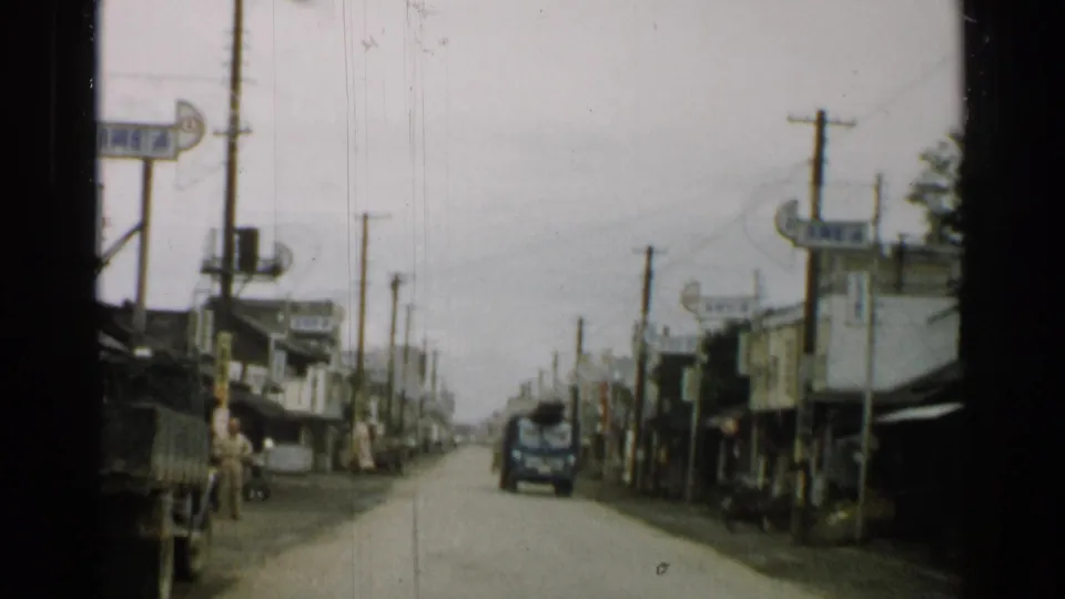A 1952 Japan 8mm frame looks down a modest town street with a vehicle ahead, roadside shops, utility poles, signs, and pedestrians near the road.