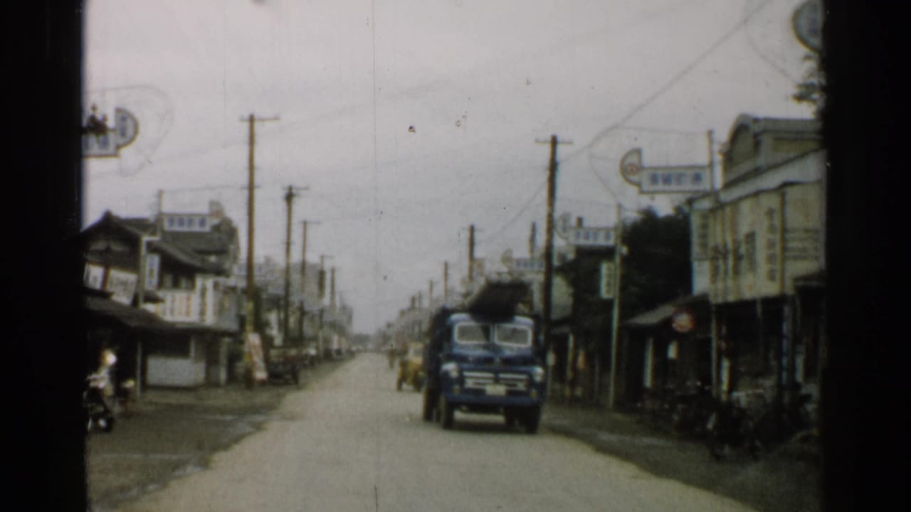 Blue truck drives along a town road lined with wooden buildings, signs, poles, and bicycles.