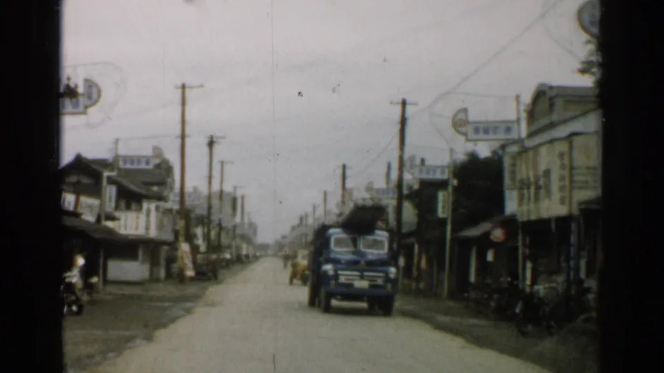 1952 Japan street view with a blue truck approaching on a road between wooden storefronts, utility poles, signs, and bicycles.