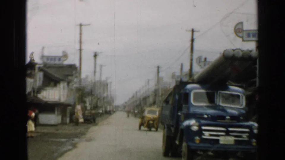 1952 Japan road scene with a blue truck close to the camera, a yellow three-wheeled vehicle, storefronts, poles, and overhead wires.