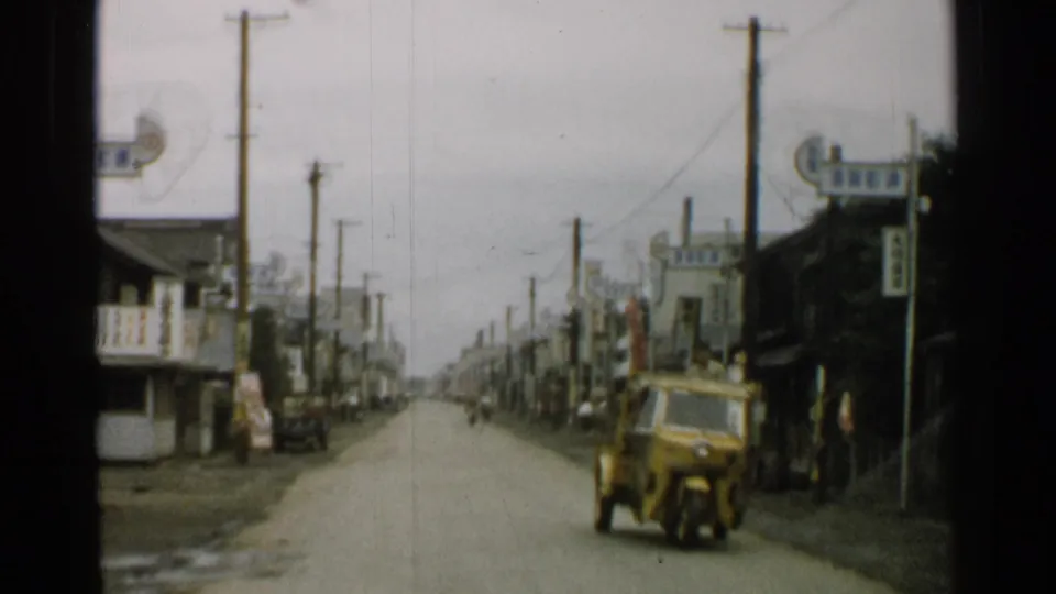 1952 Japan street scene showing a yellow three-wheeled vehicle on a road with wooden shops, signage, utility poles, and bicycles nearby.