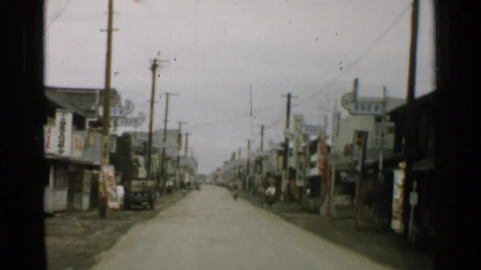 1952 Japan town road with wooden buildings, overhead wires, utility poles, signs, bicycles, and a small vehicle farther along the street.